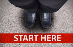 Black leather shoes seen from above, standing on asphalt in front of a red banner reading "Start here"