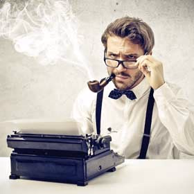 Man with glasses and a bow tie smoking a pipe behind a typewriter, looking over his glasses