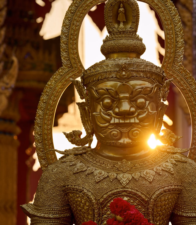 Golden guardian statue with a fierce face and ornate halo, the sun glinting behind its shoulder, red flowers at its chest