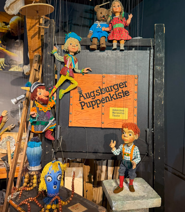 Marionettes and puppets around a sign reading Augsburger Puppenkiste in a museum display