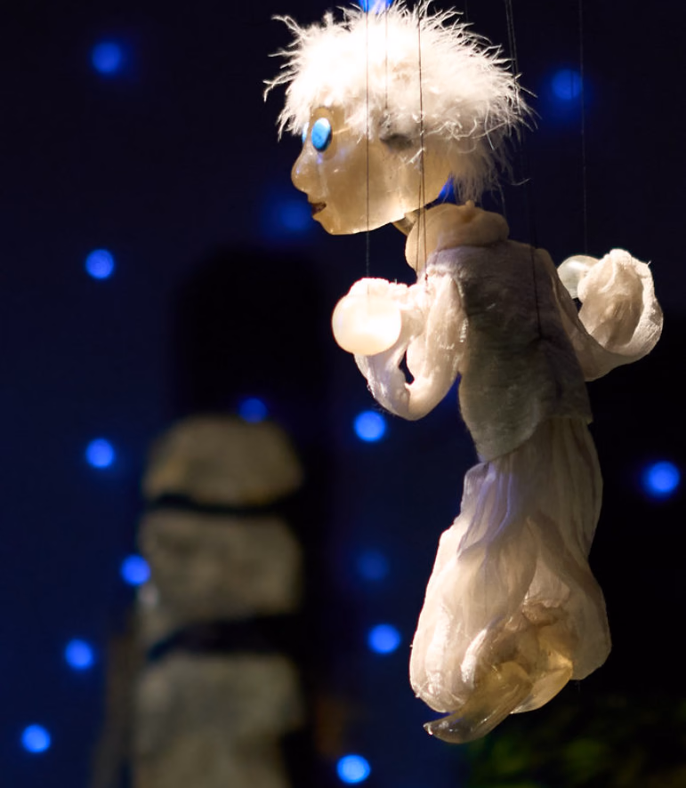 A white feathery-haired marionette with a blue eye floating against a dark blue background with glowing lights