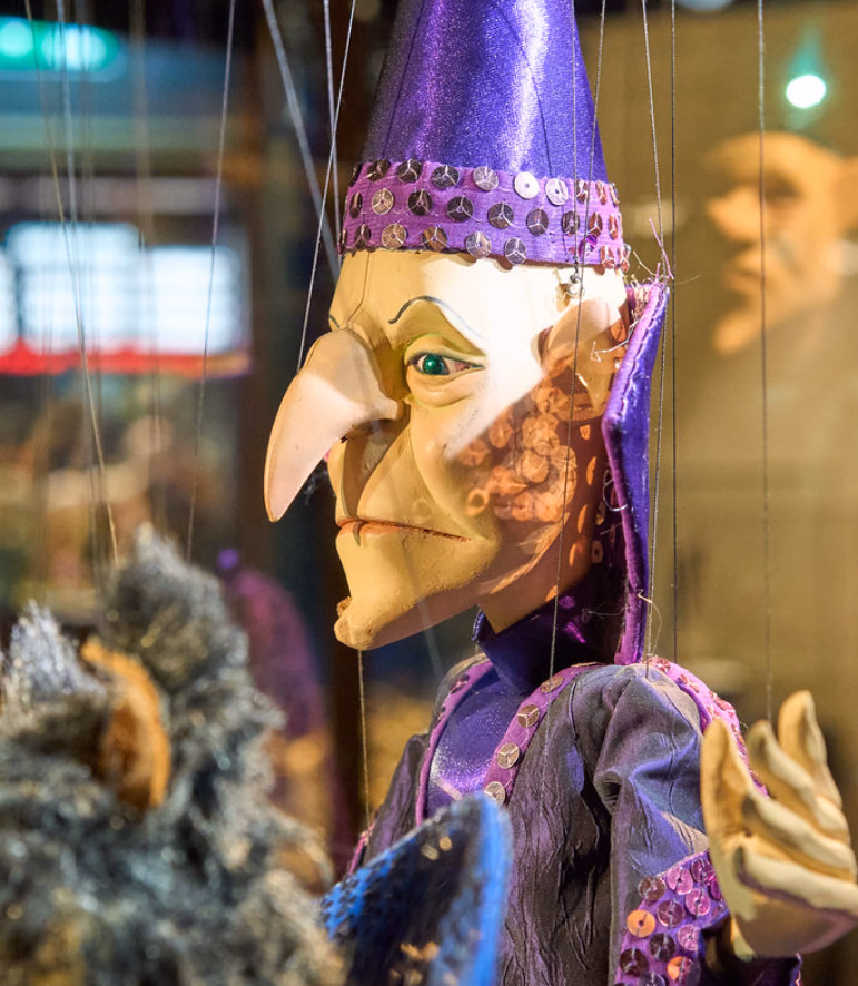 Marionette with a long pointed nose and a purple sequined pointed hat, seen behind glass