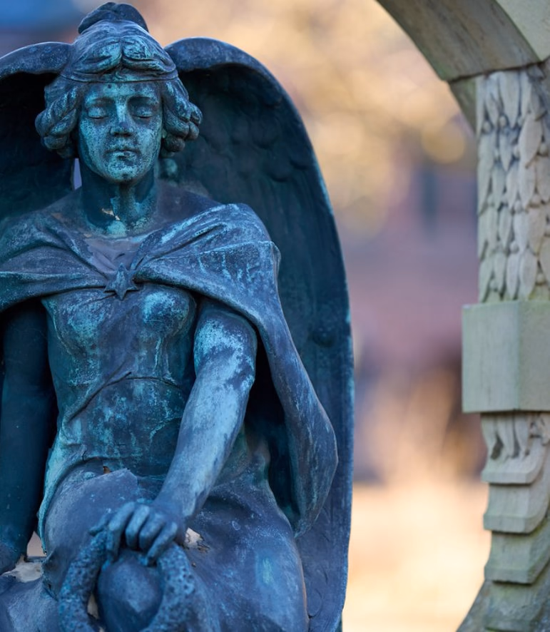 Weathered green bronze statue of a winged figure with closed eyes beside a stone arch
