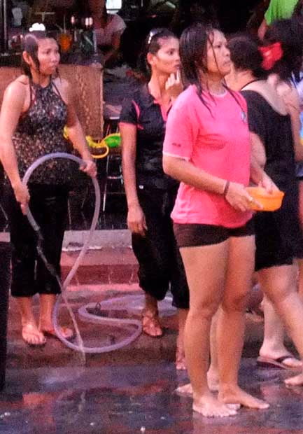 Wet women on a street during Songkran, one holding a garden hose, another a bowl, waiting for their next target