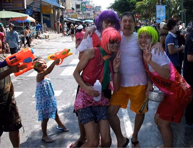 Three people in bright wigs posing with peace signs on a street during Songkran, with a child firing an orange water pistol
